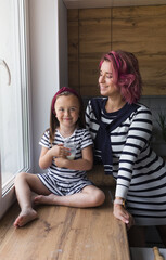 happy family, mother and her little daughter are sitting on the windowsill in the kitchen and drinking milk. They are dressed in beautiful dresses with black and white stripes