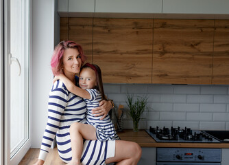 happy family, mother and her little daughter sitting on the windowsill and gently hugging each...