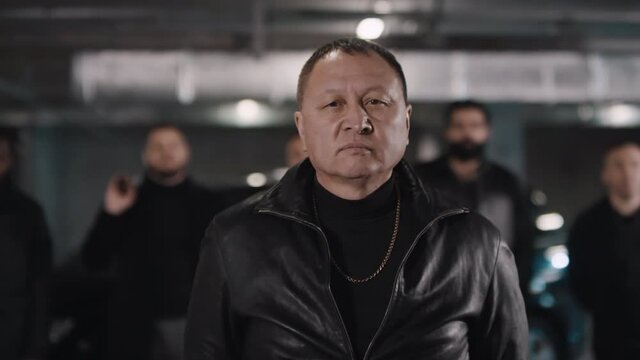 Medium closeup of assertive Asian gangster leader looking at camera ahead his armed with handguns multi ethnic gang at isolated underground parking