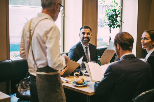 Rear view of waiter talking to business people while standing by table in restaurant