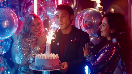 Charming guy and girls standing with cake in club. Man blowing candles at party