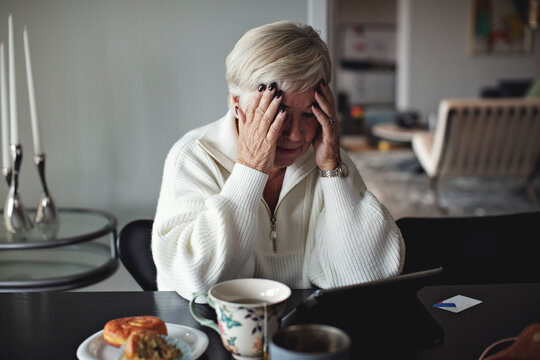 Worried senior woman with head in hands looking at digital tablet while sitting by dining table at home