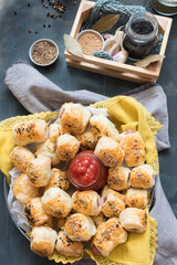 Overhead view of a metallic dish full of baked sausage rolls on a yellow cloth with a ketchup jar in the middle. A wooden basket with some spice jars inside is at the upper part of the image.