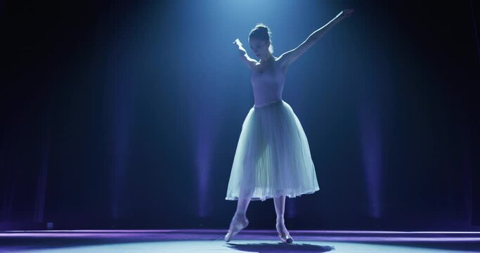 Cinematic shot of an young graceful classical ballet female dancer in white tutu is performing a choreography on classic theatre stage with spotlights before start of a show.
