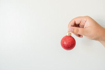 A woman's hand holding a red glittered Christmas tree ornament up against a white creamed color wall.  - 388909619