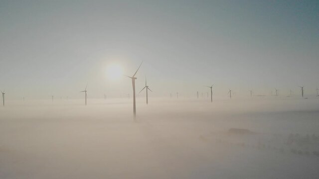 Wind farm standing tall above snow covered fields and a freezing fog on a cold winter morning.  The sun is rising behind the wind turbine casting their shadows across the fog.