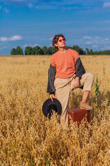 Naklejka na ściany i meble Tall handsome man with black hat and pink sunglasses standing with one leg on a brown vintage leather suitcase at golden oat field.
