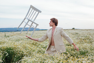 Naklejka na ściany i meble Tall handsome man standing in the middle of camomile flowers field and balancing with white chair on his palm