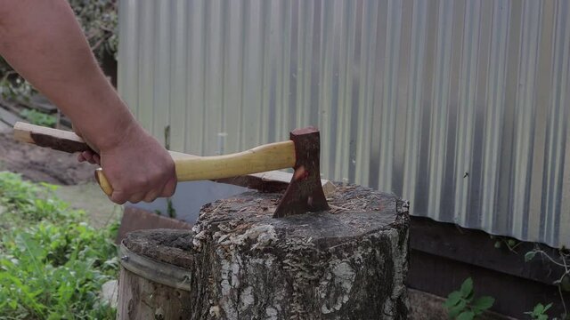 Male hands chop a stick on a stump with an ax. Procurement of firewood from woodworking waste. In the background is a corrugated sheet metal wall. Warm summer day. Side view. Soft light.