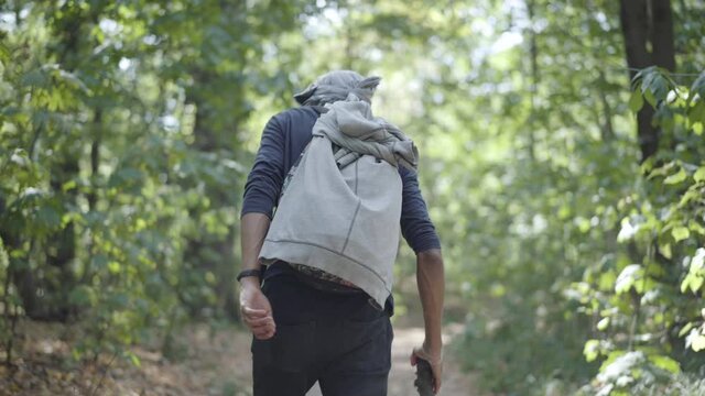 Back view of homeless escapee strolling in sunlight in summer forest. Camera follows tired young male refugee walking with wooden stick outdoors. Migration crisis concept.