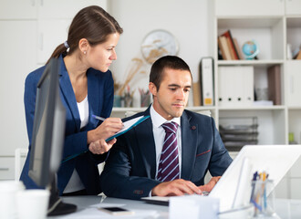 Focused young female assistant standing with clipboard near boss in office, writing down tasks