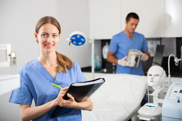 Positive female doctor cosmetologist wearing blue overall meeting client while male nurse preparing equipment before procedure