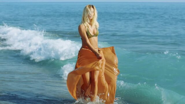 Blonde-haired female model is running by the sea in bikini and orange pareo  flowing underneath. The girl turns to the camera and poses. 2.
