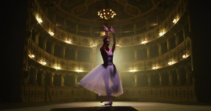 Cinematic shot of an young graceful classical ballet female dancer in white tutu is performing a choreography on classic theatre stage with dramatic lighting before start of a show.
