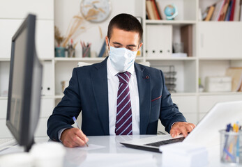 Focused businessman in disposable face mask working on laptop in office. Necessary precautions during COVID 19 pandemic..