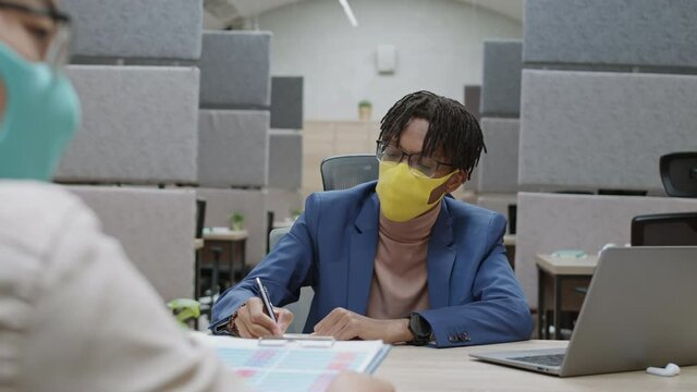 Rear view of young African businessman wearing medical mask sitting at desktop in modern office, having meeting with colleagues and then looking at camera