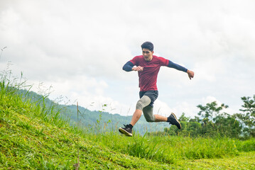 Healthy handsome Asian man in sportswear running on the mountain trail in summer day. Strong athlete male enjoy jogging on grass hill. Outdoor workout lifestyle and extreme sport training concept.