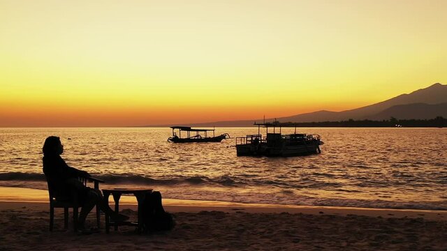 Silhouette Of A Woman Sitting On A Wooden Chair Facing Rightwards On The Beach As She Relaxes Nearby The Wide Waving Sea With Boats Floating Still During The Afternoon, Slowly Tracking Forward.