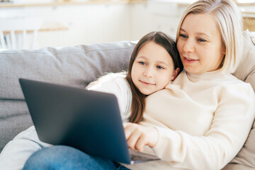 Caucasian mommy with her daughter lying on the couch talking and looking at the laptop screen. Home...