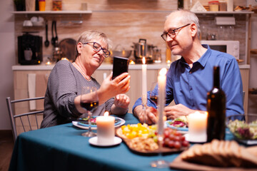Senior wife showing photos on phone during relationship anniversary in the evening. Sitting at the table in the kitchen, browsing, searching, using phone, internet, celebrating their anniversary.