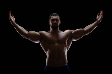 Fototapeta na wymiar Portrait of a bodybuilder standing isolated on black background in a shadow with raised hands to show off his muscles
