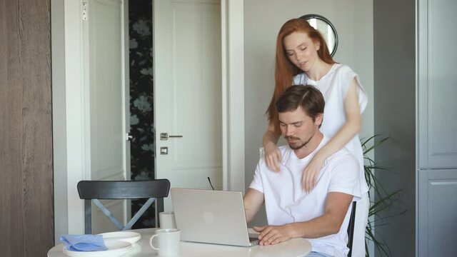 Redhead woman gives massage to her working husband, young lady take care of bearded man sitting with laptop
