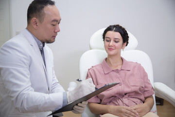 Fototapeta premium Young pretty woman in beauty salon consults with beautician on anti-aging treatments. Doctor writing notes on clipboard and explain procedures to the patient