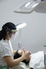 Relaxed woman lying in salon and having deep cleansing nourishing facial treatment. Beautician wearing visor and applying cream and doing face massage to client face. Healthcare, wellness and medicine