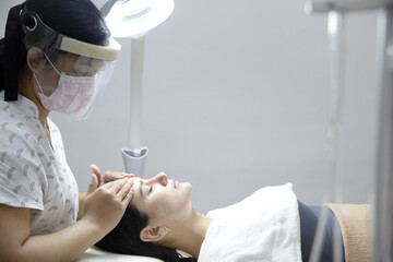 Relaxed woman lying in salon and having deep cleansing nourishing facial treatment. Beautician wearing visor and applying cream and doing face massage to client face. Healthcare, wellness and medicine