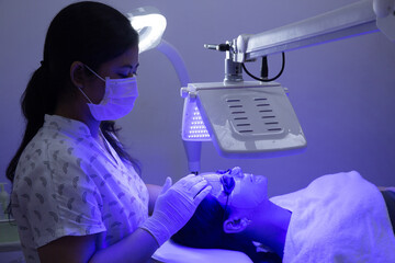Young woman having blue LED light facial therapy treatment in beauty salon. Beautician wearing face...