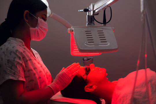 Young woman having red LED light facial therapy treatment in beauty salon. Beautician wearing face mask maintaining safety procedures during appointment. Beauty, new normal and wellness