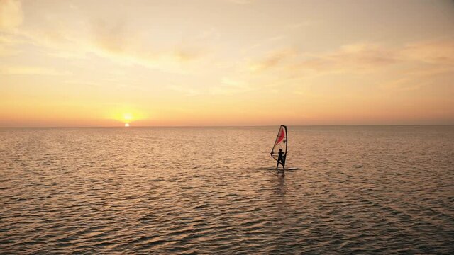 Distant silhouette of person sailing contemporary windsurf board training on calm ocean at sunset at tropical resort