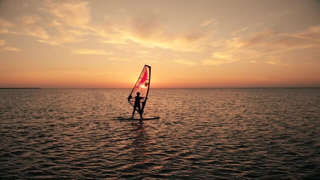 Dark silhouette of woman sailing contemporary windsurf board training on calm ocean water against sunset at resort zoom out