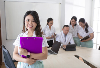 Young asian female nurse, student or practitioner standing in front of medical team holding clipboard looking at camera and smiling. Group of people behind discussing and interacting with each other