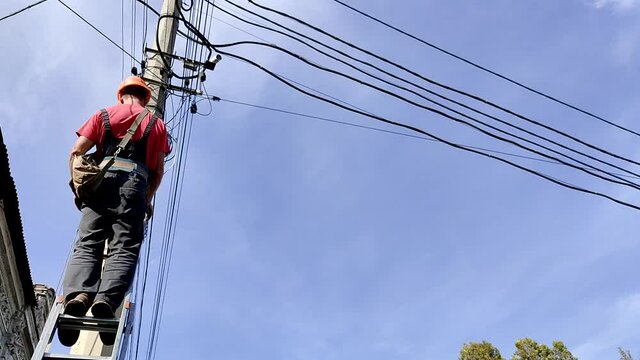 Skilled electrician in helmet fixes wires standing on ladder near high pole against blue sky on summer day backside view