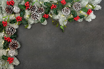 Festive winter solstice snow covered spruce fir border with holly, mistletoe & pine cones on grey grunge. Composition for Christmas & New Year holiday season. Top view, flat lay, copy space.