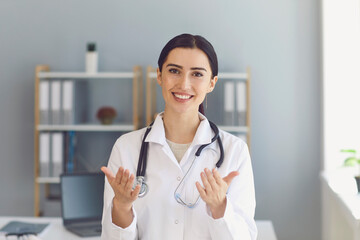Female doctor giving medical consultation to patient online from clinic. Young GP communicating to...
