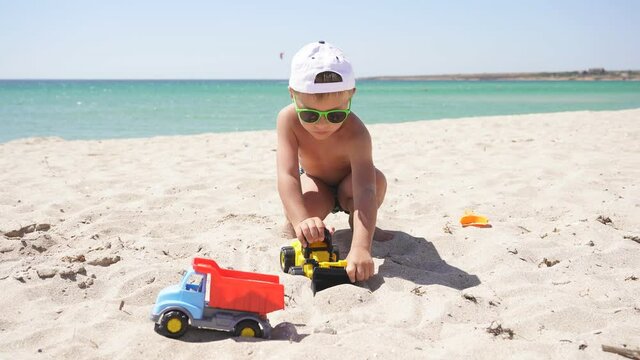 Tanned boy playing with cars on the beach, plastic toy car, excavator.