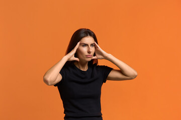 Leave me alone. Tired gloomy caucasian woman in black t-shirt holds hands on temples, has headache, feels exhausted and tension, looks sideways. Isolated over an orange background.