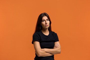 Can you find a solution. Confused looking caucasian woman in black t-shirt holds hands crossed, looks hopefully at camera, asks for help. Isolated over an orange background.