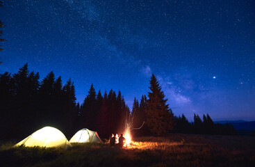 Evening camping with campfire. People having a rest near tent city, enjoying valley of mountains in pine forest. Dark blue night sky is strewn with bright stars and Milky Way is visible on it.