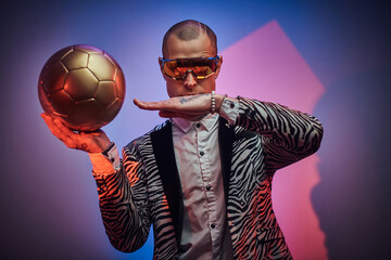Handsome guy dressed in custom suit with short haircut and eyewears poses with golden soccer ball...