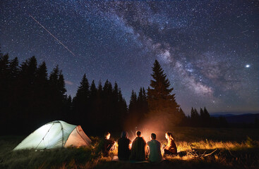 Abendliches Sommercamping, Fichtenwald im Hintergrund, Himmel mit Sternschnuppen und Milchstraße. Eine Gruppe von fünf Freunden sitzt zusammen am Lagerfeuer in den Bergen und genießt die frische Luft in der Nähe des beleuchteten Zeltes