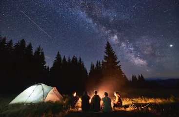 Foto auf Acrylglas Camping Abendliches Sommercamping, Fichtenwald im Hintergrund, Himmel mit Sternschnuppen und Milchstraße. Eine Gruppe von fünf Freunden sitzt zusammen am Lagerfeuer in den Bergen und genießt die frische Luft in der Nähe des beleuchteten Zeltes