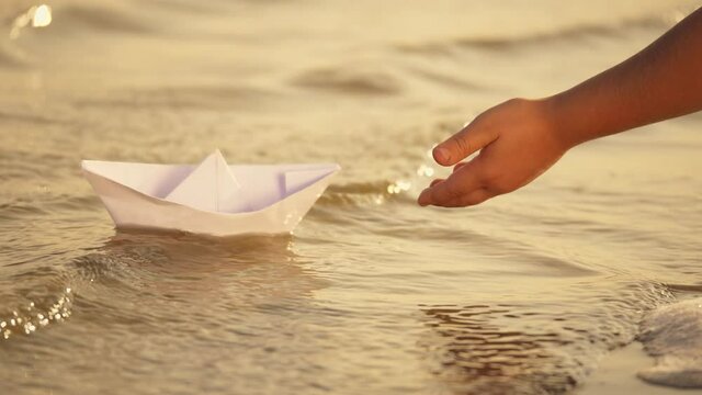 Hand of a little girl lowers a paper boat into the river. Children's paper boat on the waves of the river. Close-up of a little girl's hand with a paper boat
