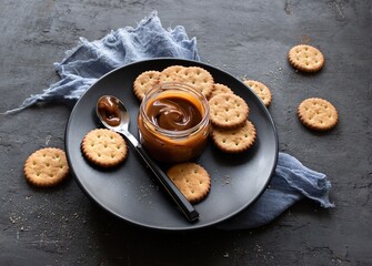 Salted caramel jar with a spoon and crunchy cracker on a black plate on a dark background. Top view 