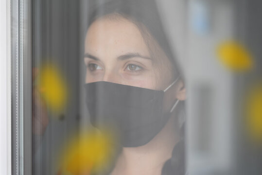 Young Caucasian Teenage Girl Wearing Protective Face Mask, In Nursing Care Home, Looking Outside Window With Sadness In Her Eyes, Self Isolation Due To The Global COVID-19 Coronavirus Pandemic