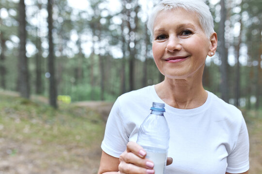 Close up image of attractive short haired middle aged woman in white t-shirt posing outdoors with pines in background, drining water from plastic bottle, having break during cardio workout
