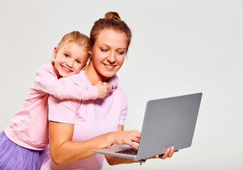 Closeup portrait of сute girl 4-5 year old and her mother in pink clothes, using laptop on gray background. Free space for advertisement.