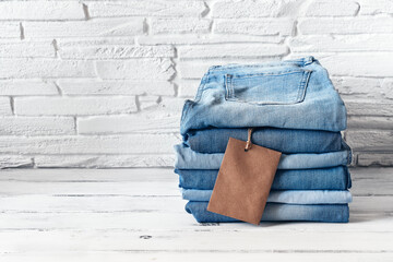A stack of blue jeans with a brown blank tag on a white wooden table, brick backing. Heap of stylish trendy denim pants. Selective focus.